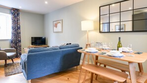 Uma sala de estar moderna com um sofá azul, mesa de jantar de madeira para quatro pessoas e uma televisão no canto.