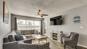 A living room with a sectional sofa, a coffee table, a patterned armchair, a ceiling fan, and a flat-screen TV mounted on the wall. - Explore Boardwalk Thrills! Central Destin Condo (Destin)