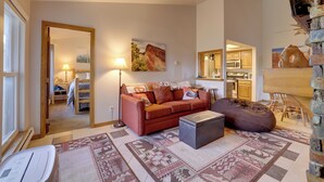 Ein Wohnzimmer mit Sofa, Hocker und Teppich. Im Hintergrund ist eine Küchenzeile erkennbar.