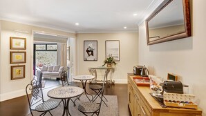 Un salon bien éclairé, avec un canapé, une table basse et un coin repas comprenant des chaises. On y trouve des tableaux encadrés accrochés au mur, une plante en pot et un meuble en bois abritant une machine à café.