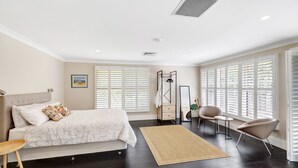 Une chambre à coucher comprenant un lit, une table de chevet, une chaise et de grandes fenêtres équipées de stores.