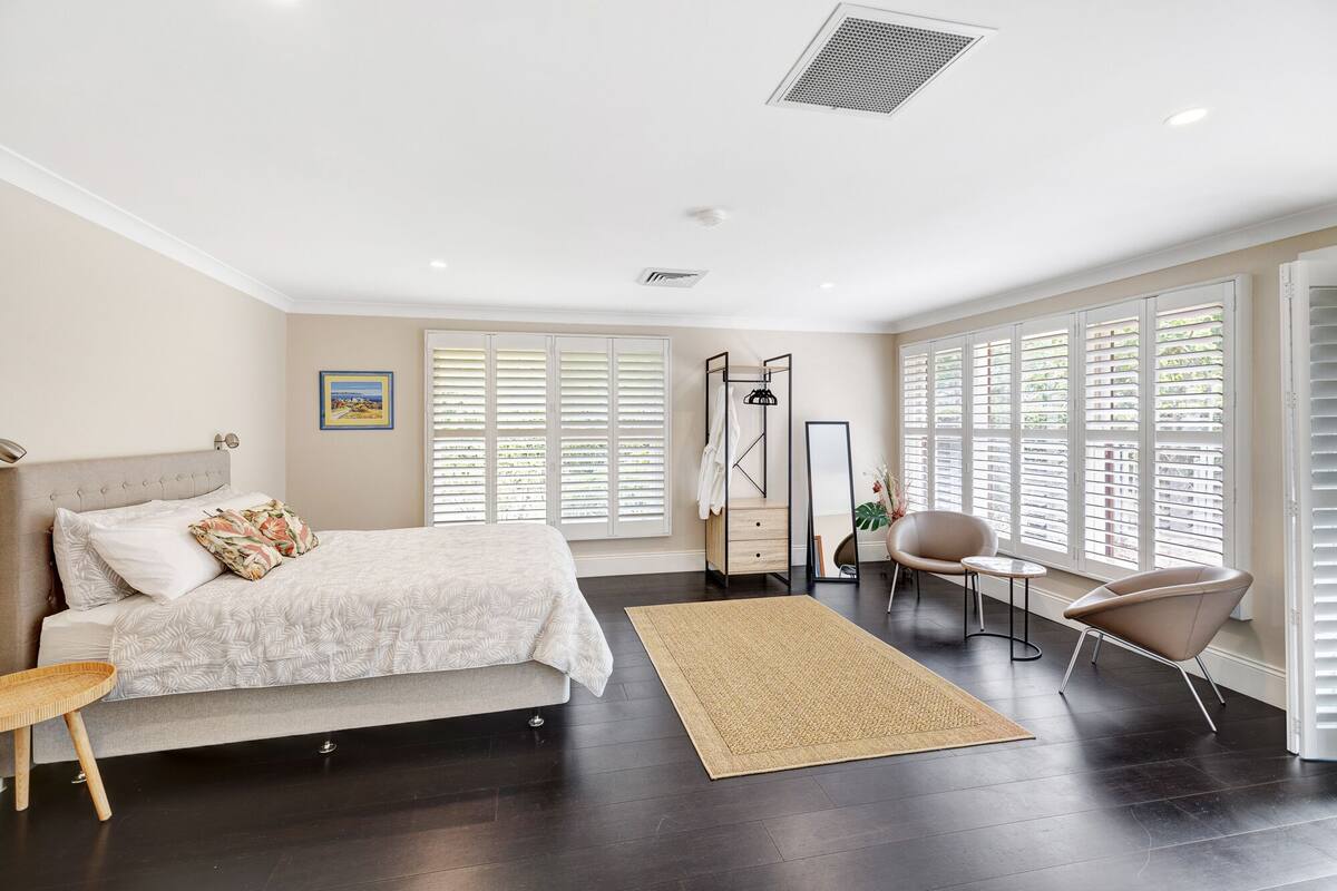 A bedroom with a bed, bedside table, chair, and large windows with blinds.