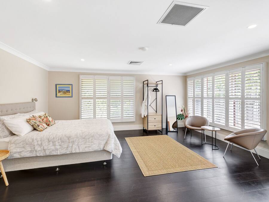 A bedroom with a bed, bedside table, chair, and large windows with blinds.