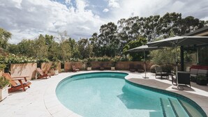 Une piscine incurvée, dont le bord est recouvert de carreaux, entourée d’une terrasse avec des chaises en bois et un espace salon abrité.