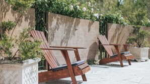 Deux fauteuils de jardin en bois, dotés d’une structure en métal, sont placés sur une terrasse carrelée. Un mur en pierre et de la verdure se trouvent en arrière-plan.