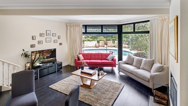 Un salon spacieux avec un canapé rouge, un canapé beige et un téléviseur posé sur un meuble noir. On peut apercevoir une piscine à travers les grandes fenêtres.