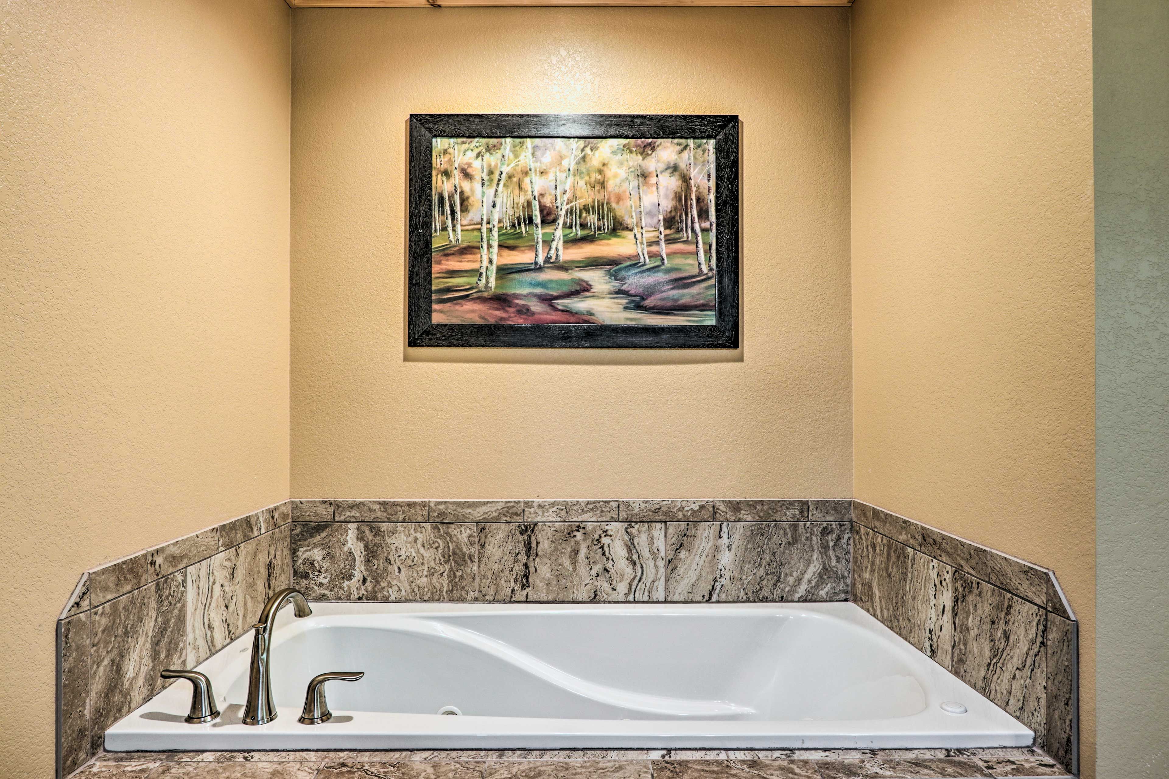 A bathroom with a marble bathtub, a framed painting of a forest scene, and a beige wall.