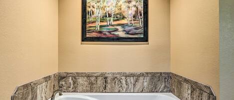 A bathroom with a marble bathtub, a framed painting of a forest scene, and a beige wall.
