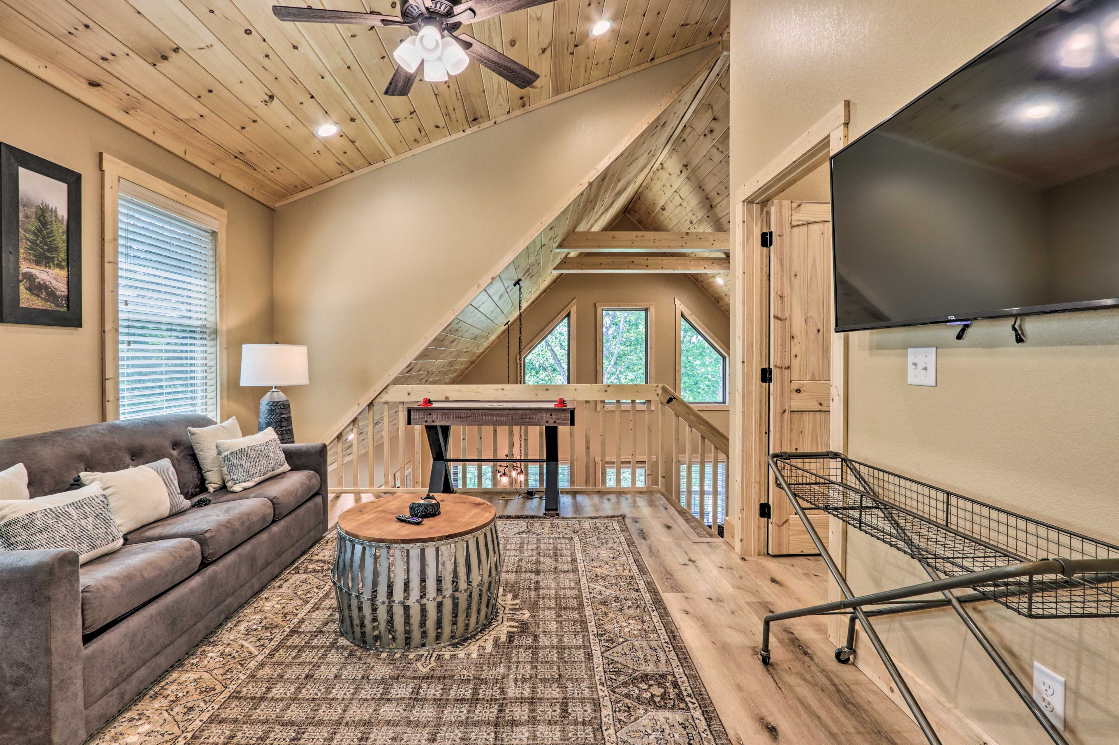 A cozy loft-style room with a wooden ceiling, a large window, a sofa, a coffee table, and a TV.