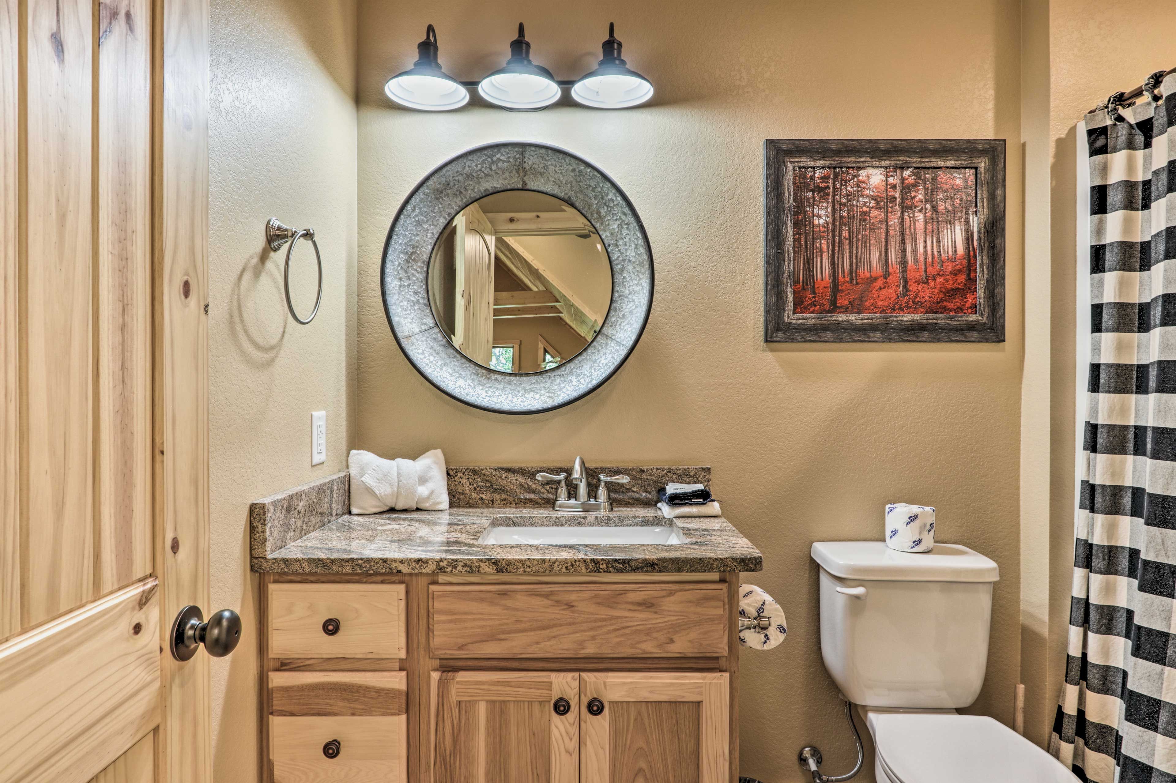A bathroom with a wooden vanity, a round mirror, a sink, a toilet, and a framed picture of a forest.