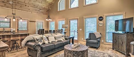 A spacious living room with a high ceiling, wooden beams, and large windows. There is a dark-colored sofa with multiple cushions, a round ottoman, and a television on a wooden cabinet.