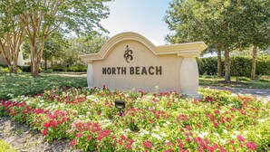 Een bord met de tekst 'NORTH BEACH', met een decoratieve boog en een tuin vol roze en witte bloemen er voor.