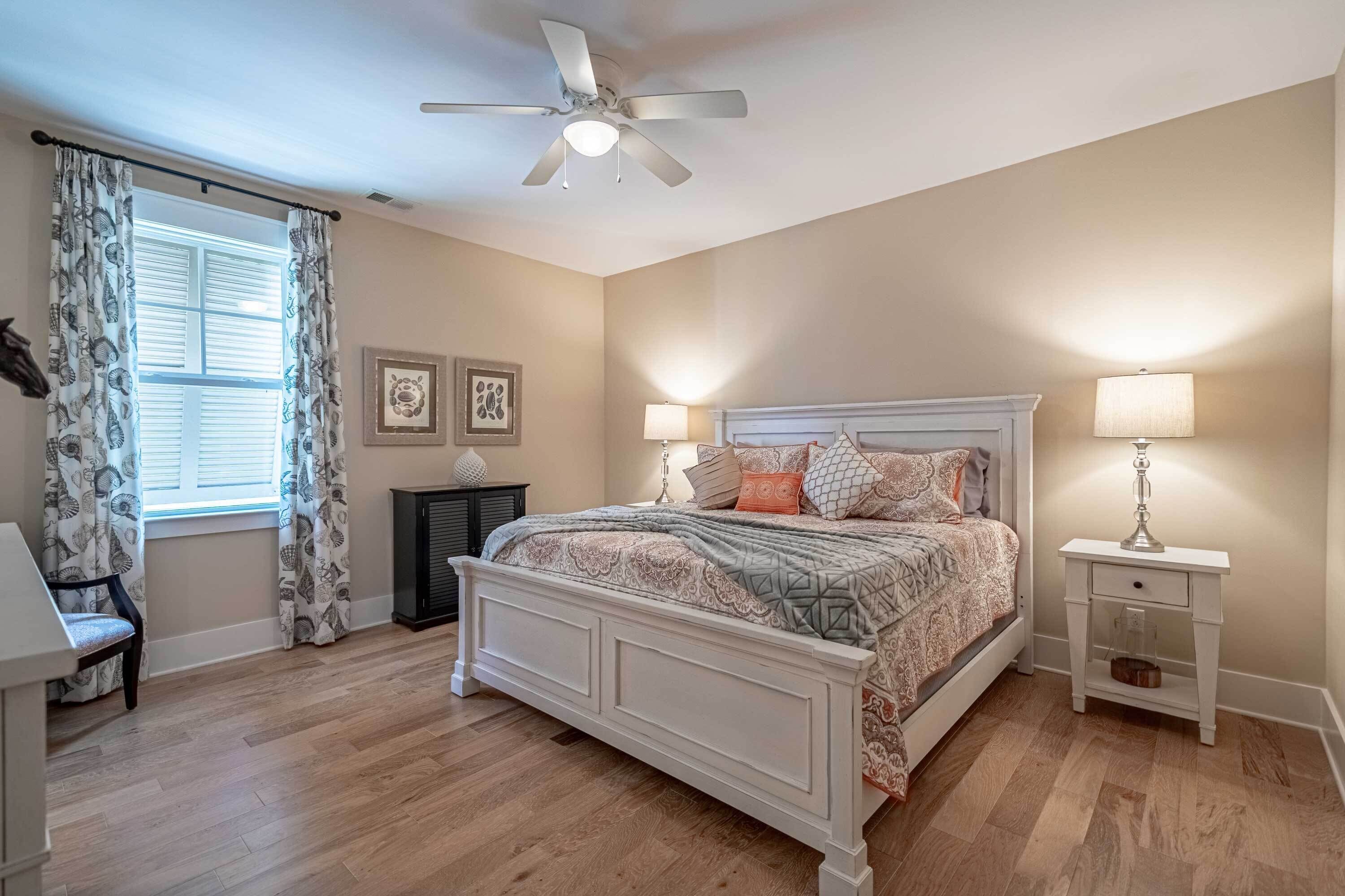 A bedroom with a bed, nightstands, a ceiling fan, a window with curtains, and a wall with framed pictures.