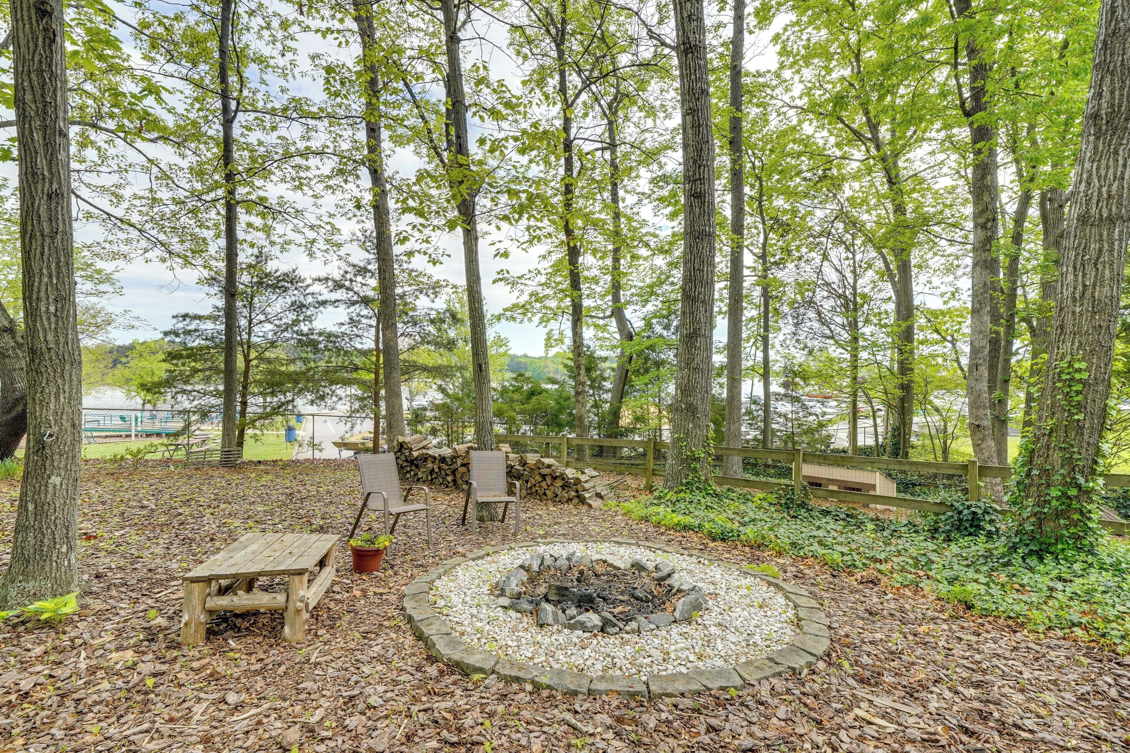 A forested area with a circular stone fire pit surrounded by rocks, wooden benches, and a picnic table.