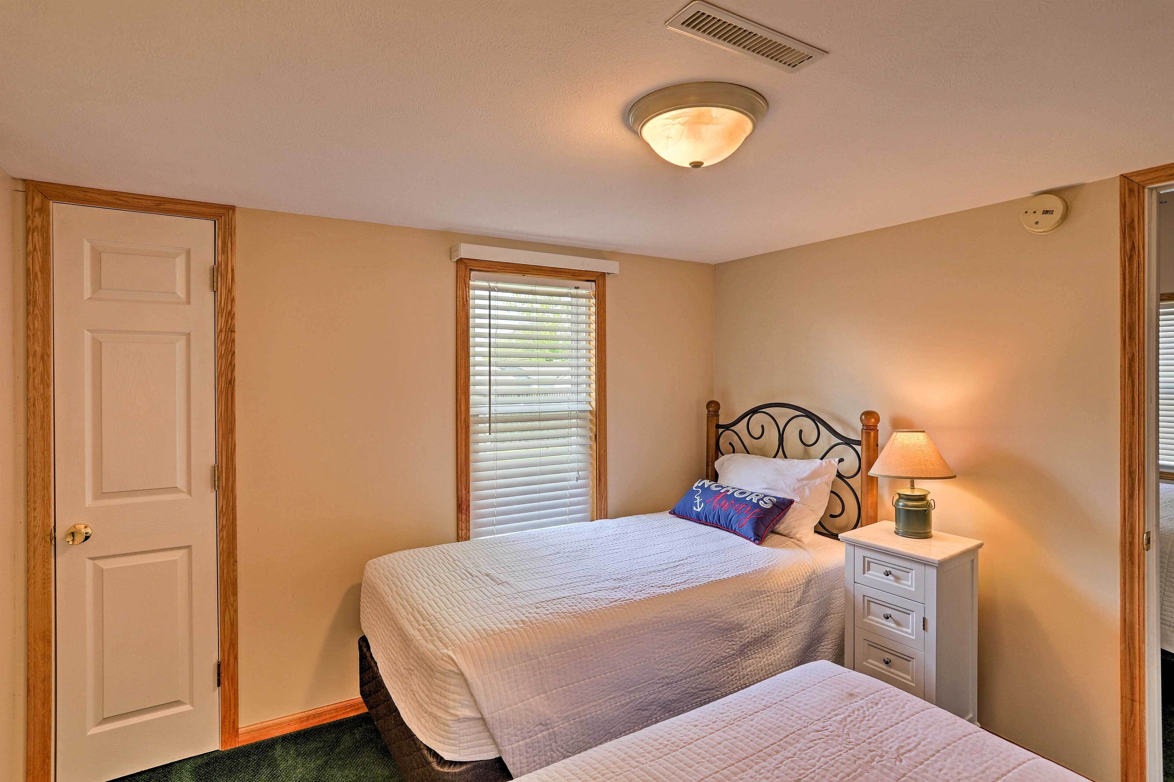 A bedroom with two beds, a window with blinds, a bedside table with a lamp, and a door.