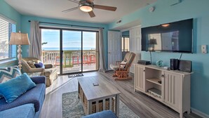 A living room with a ceiling fan, a TV, and a view of the ocean through large sliding doors. - Walk to Shore! Ocean-view Condo in Carolina Beach (Carolina Beach)