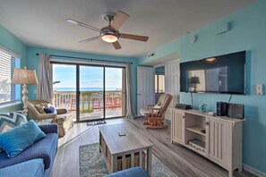 A living room with a ceiling fan, a TV, and a view of the ocean through large sliding doors. - Walk to Shore! Ocean-view Condo in Carolina Beach (Carolina Beach)