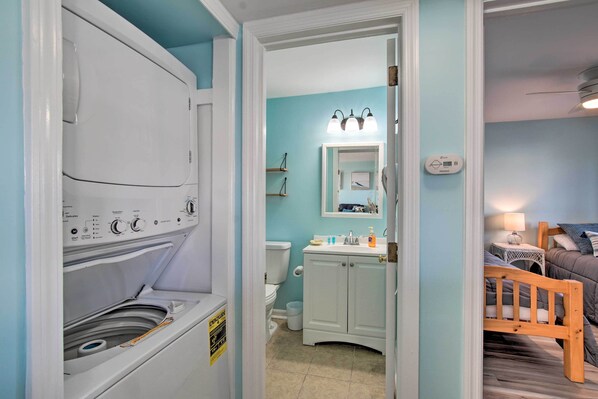 A compact bathroom with a washing machine, a toilet, a sink, and a bed in the adjacent room. - Walk to Shore! Ocean-view Condo in Carolina Beach (Carolina Beach)