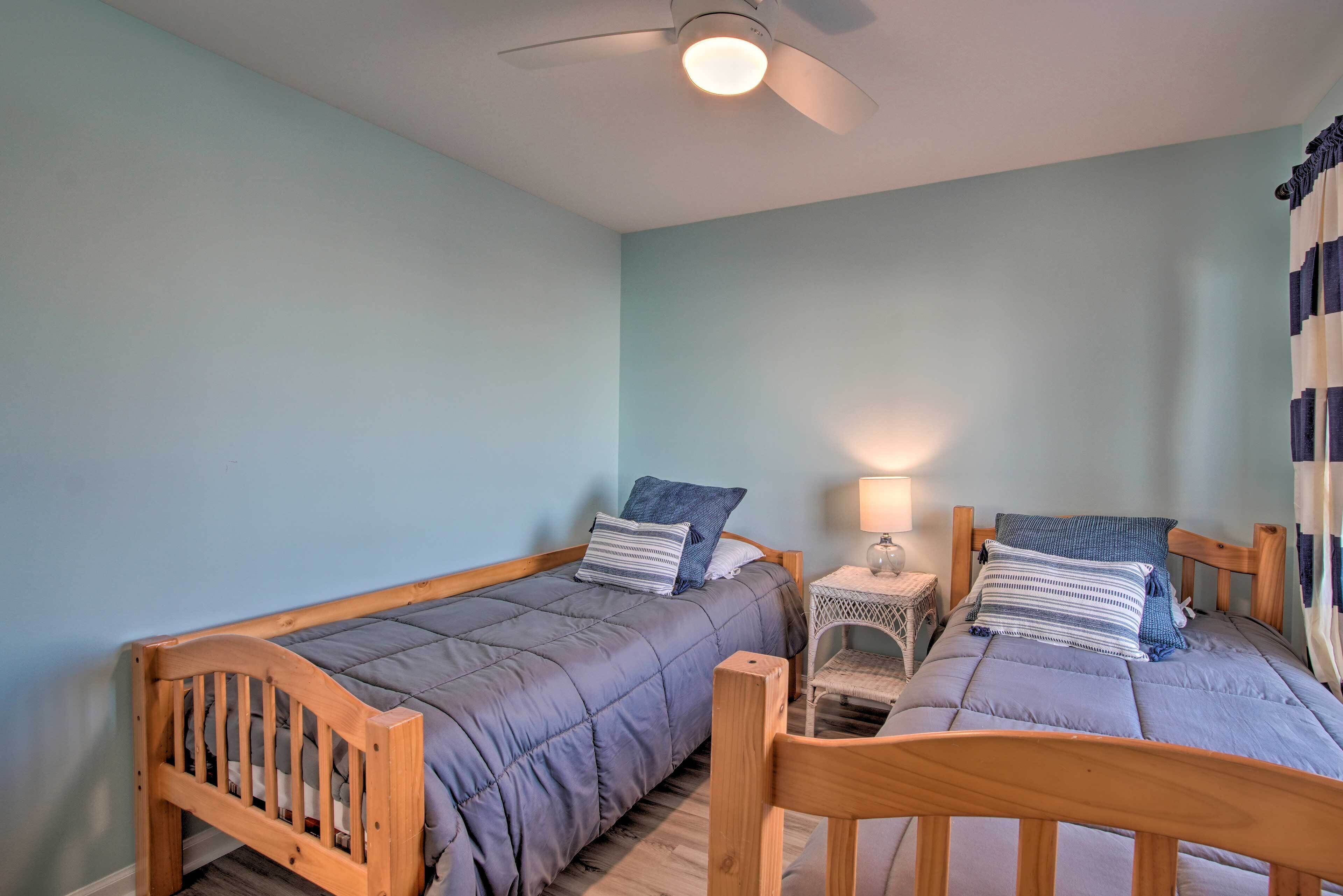 Una habitación con dos camas de madera, cada una con un edredón y almohadas grises, una mesita de noche blanca pequeña con una lámpara y un ventilador de techo.