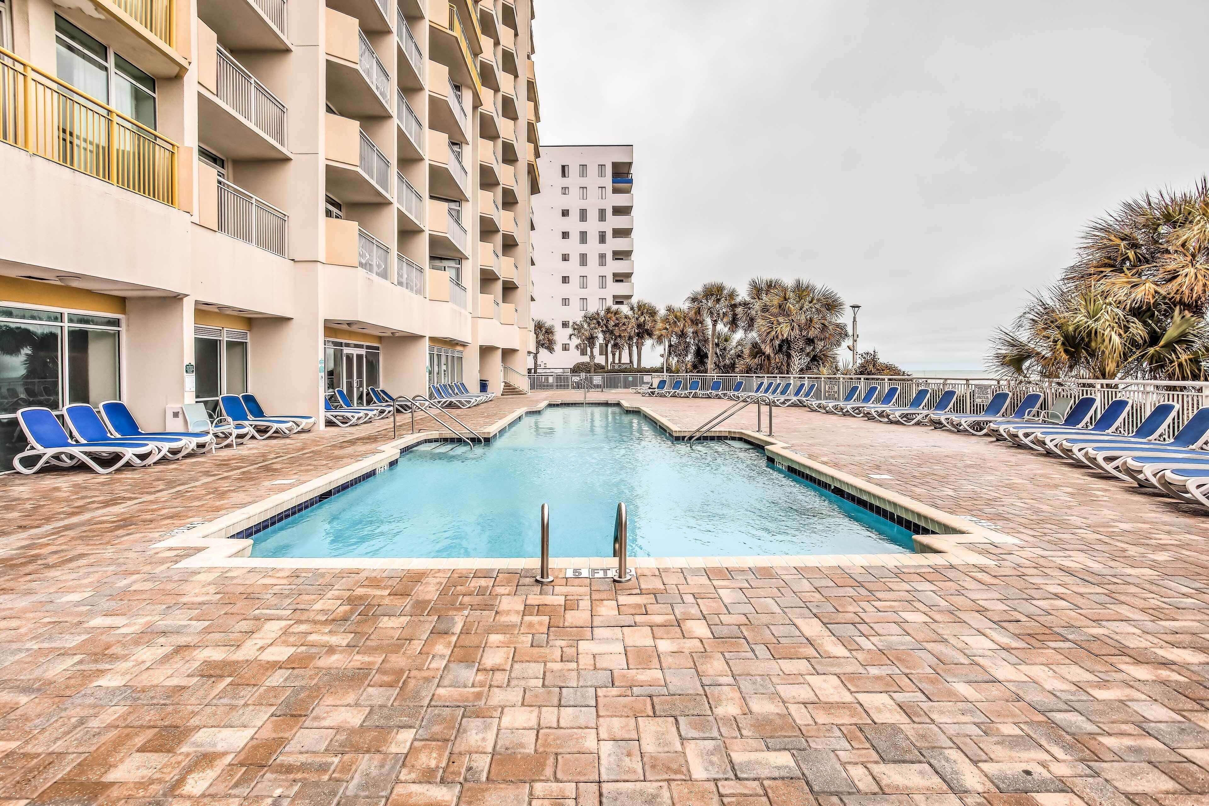 Une piscine entourée de chaises longues bleues et dotée d’un sol en briques, le tout bordé d’un immeuble de grande hauteur avec des balcons.