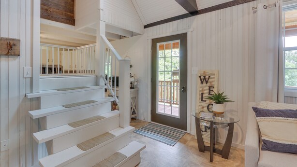 A staircase with white railings leading to a landing, a door with a 'WELCOME' sign, a small table with a plant, and a cushioned bench.
