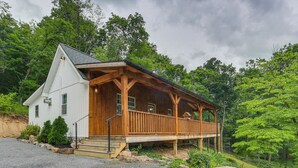 A wooden cabin with a porch, surrounded by trees and a gravel driveway.