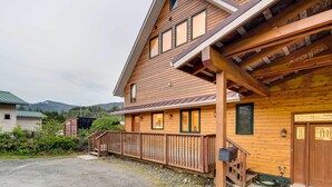 A wooden cabin with a porch, a staircase, and a mountain in the background. - Ketchikan Apt - Walk to Refuge Cove Beach! (Ketchikan)