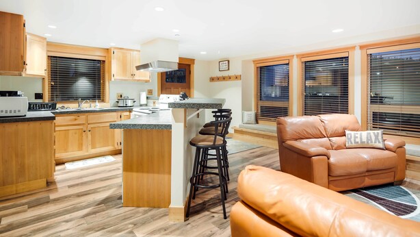 A modern kitchen with wooden cabinets, a central island, and bar stools. There is a leather sofa with a 'RELAX' pillow and a window with blinds. - Ketchikan Apt - Walk to Refuge Cove Beach! (Ketchikan)