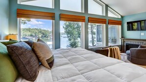 A bedroom with a large bed, multiple windows, a fireplace, and a view of a lake.