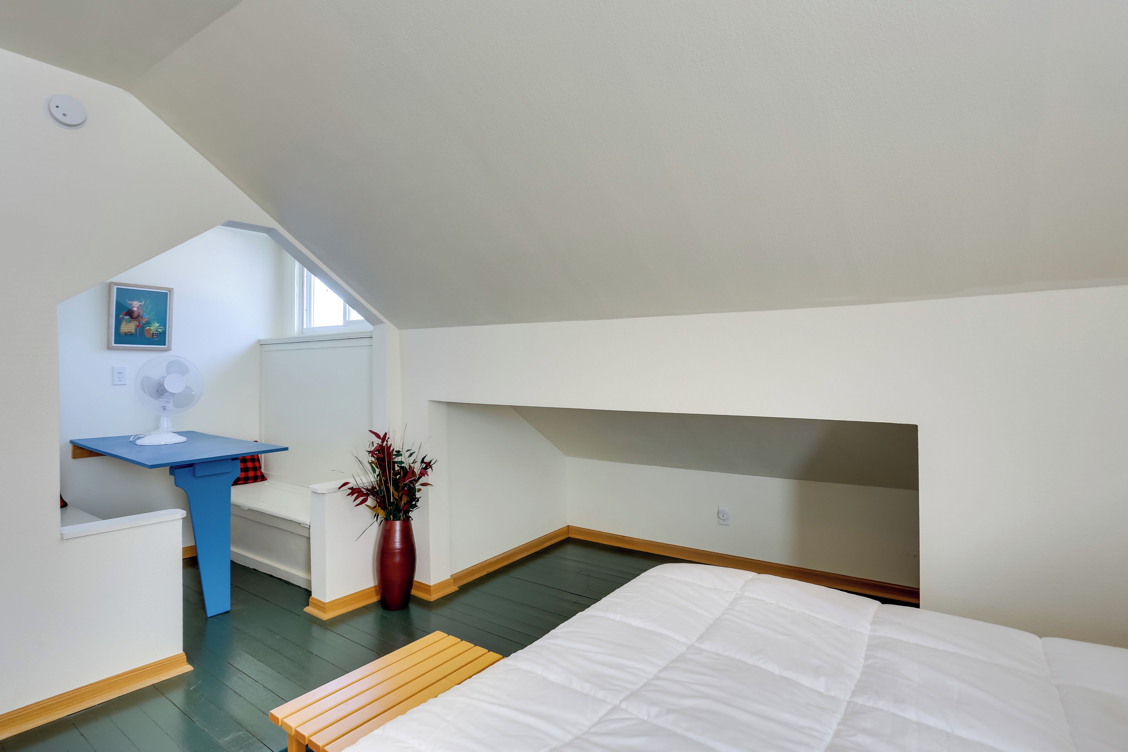 Un dormitorio con techo inclinado, cama, escritorio, silla, ventilador y un florero con flores.
