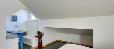 Un dormitorio con techo inclinado, cama, escritorio, silla, ventilador y un florero con flores.
