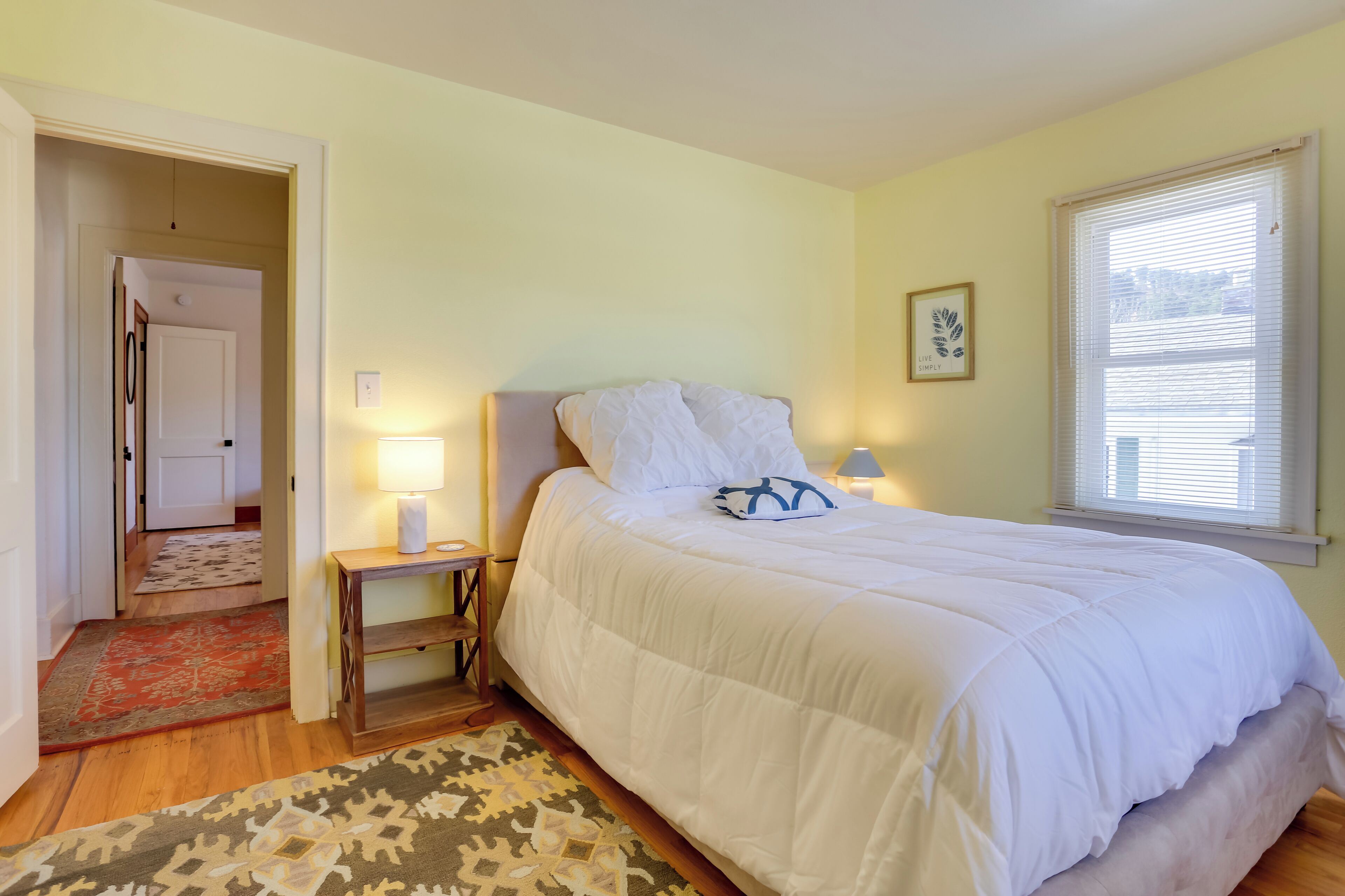 A bedroom with a large bed, a nightstand with a lamp, a window with blinds, and a door leading to another room.