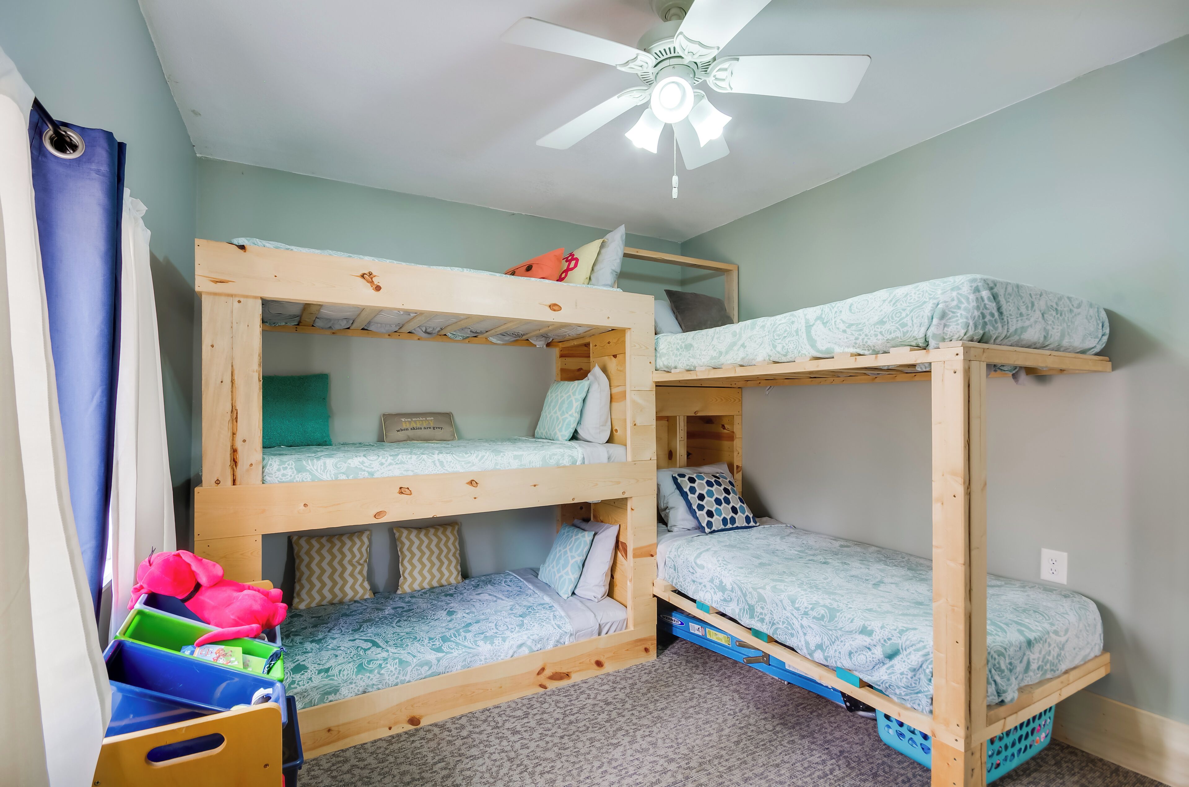A bunk bed room with a ceiling fan, a window with curtains, and a storage bin with toys.