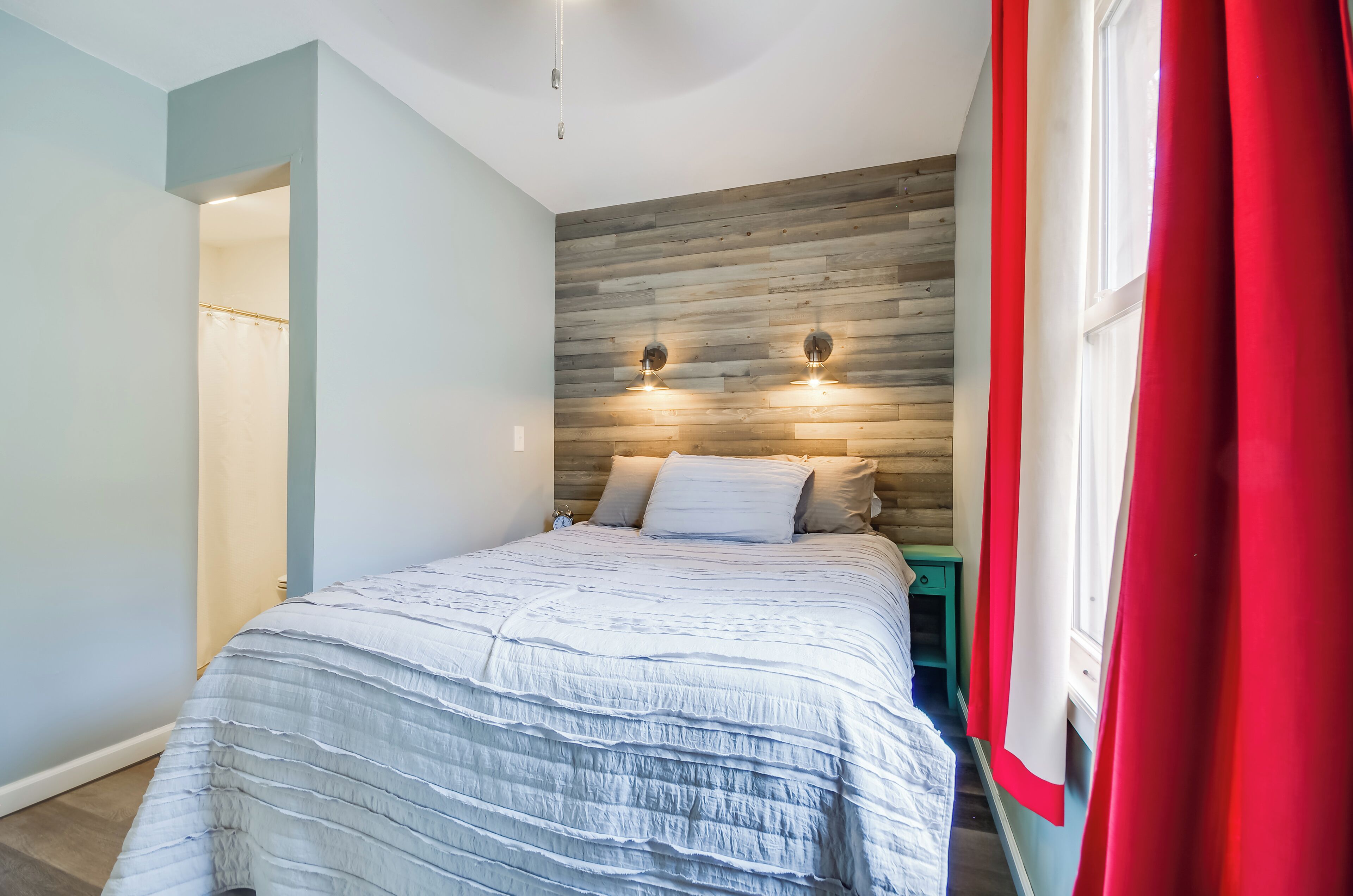 Un dormitorio con cama, cabecero de madera, dos luces fijadas a la pared, una mesita de noche y una cortina roja.