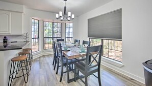 Un espace repas avec une table en bois, des chaises noires et un lustre. De grandes fenêtres offrent une vue sur l’extérieur.