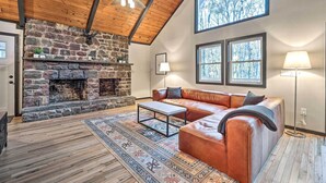 A cozy living room with a stone fireplace, leather sofa, and large windows. - Game Room, Fire Pit & Deck: Spacious Poconos Home! (Lake Harmony)