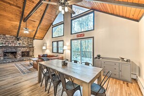 A spacious loft with a high ceiling, exposed wooden beams, a stone fireplace, a dining area with a long table and chairs, and large windows offering views of the outdoors. - Game Room, Fire Pit & Deck: Spacious Poconos Home! (Lake Harmony)