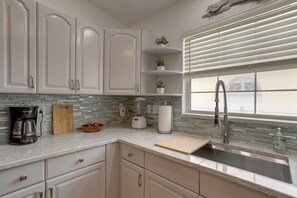 Una cocina con armarios blancos, estufa integrada, cafetera, licuadora y ventana con persianas.