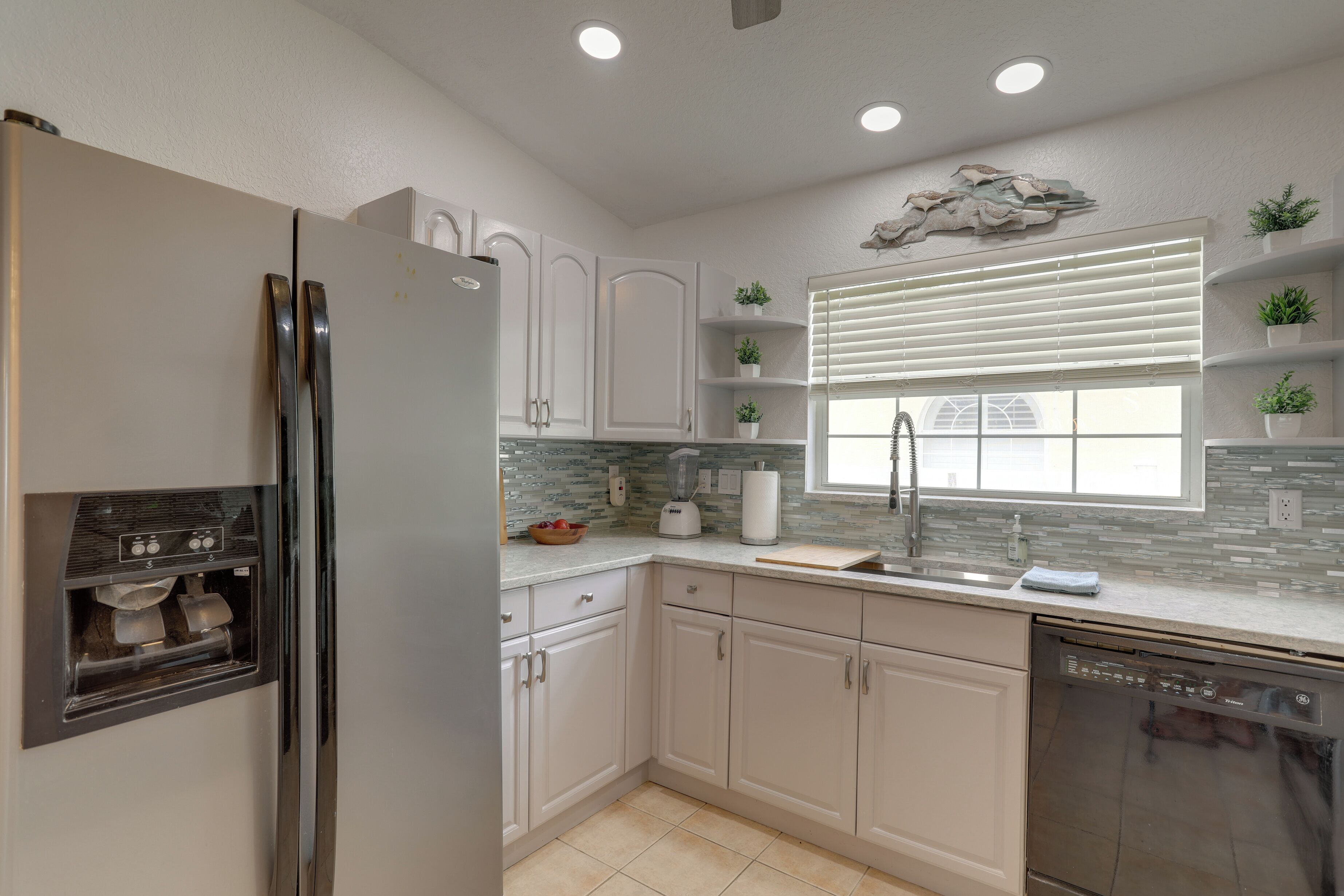Una cocina moderna con armarios blancos, refrigerador empotrado, lavavajillas y ventana con persianas.