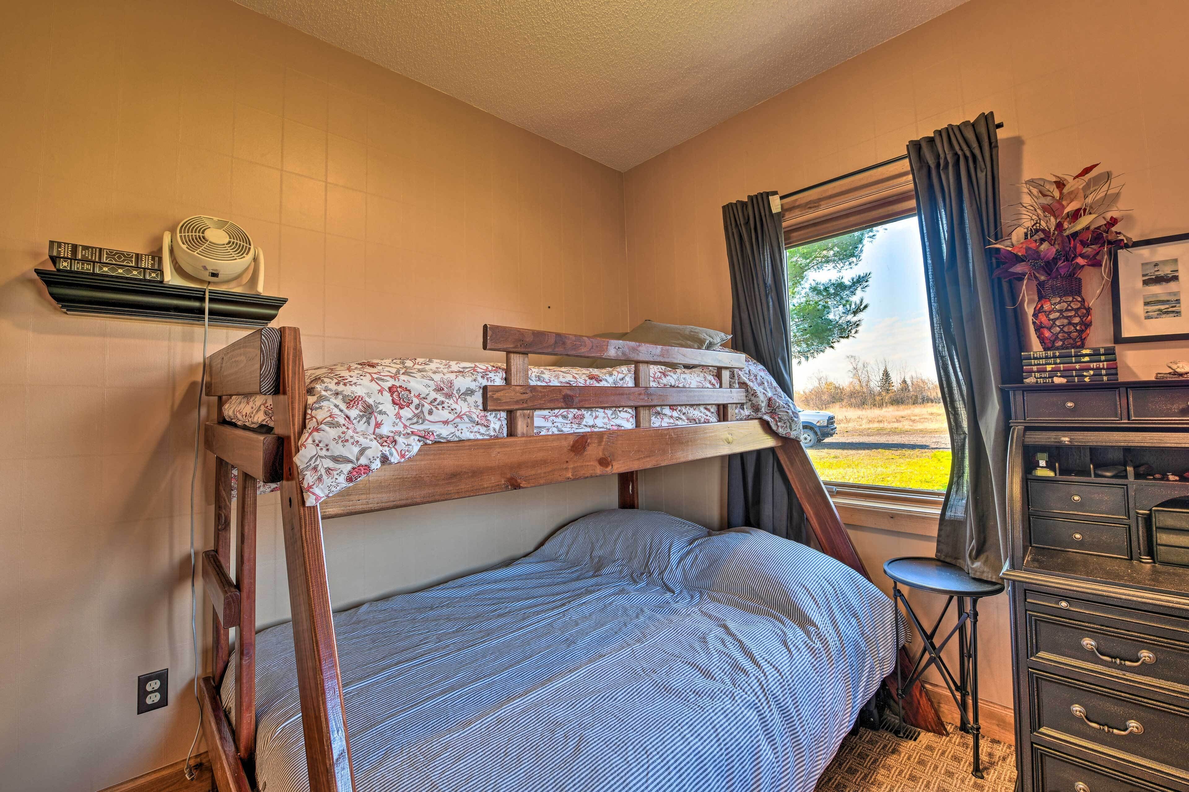A bunk bed with a floral bedspread, a small stool, a dresser with drawers, a fan on a shelf, and a window with curtains.
