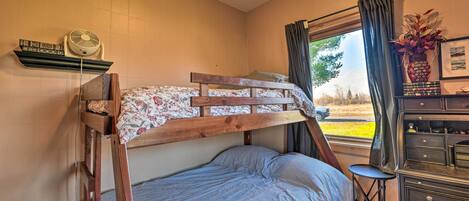 A bunk bed with a floral bedspread, a small stool, a dresser with drawers, a fan on a shelf, and a window with curtains.