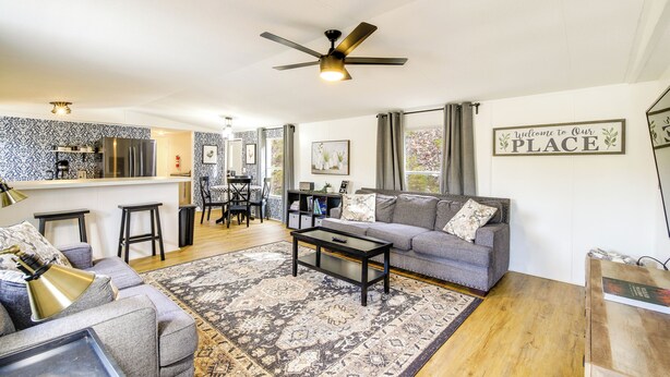A modern living room with a grey sofa, a coffee table, and a patterned rug. There is a kitchen area with a bar and a ceiling fan. - Camp-style Cabin Near Pigeon Forge: Fire Pit, Deck (Sevierville)