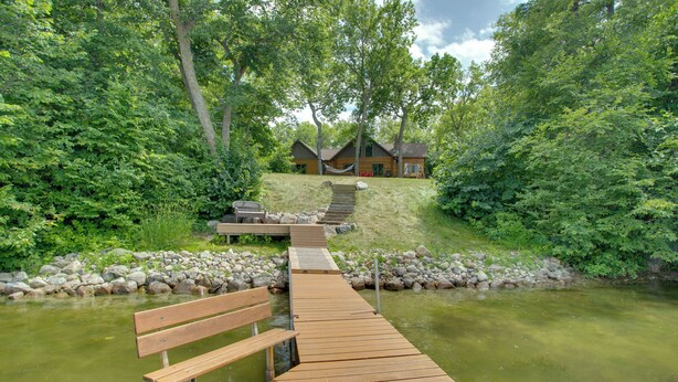A wooden dock leading to a cabin surrounded by lush greenery and a calm river.
