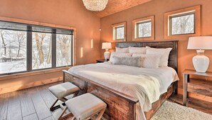 A bedroom with a large bed, wooden headboard, and a chandelier. There are three windows with views of trees and a snowy landscape.