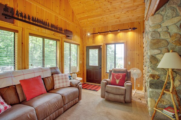 Interior acogedor de una cabaña con techo de madera, pared de piedra, un sofá de cuero, un sillón y un ventilador.