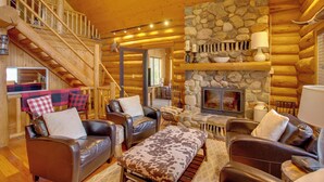 A log cabin interior with a stone fireplace, leather furniture, and a staircase leading to an upper level.
