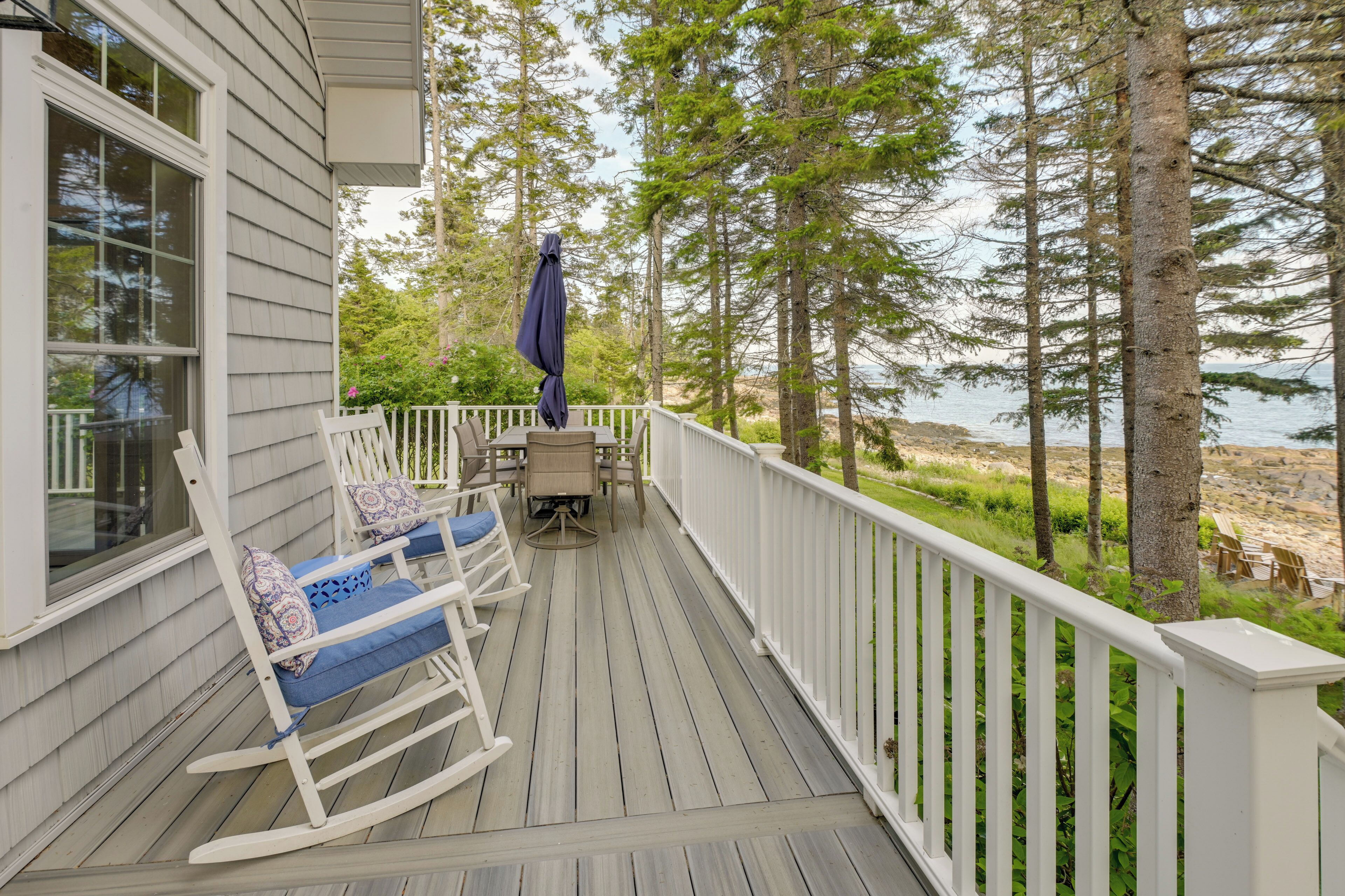 A wooden deck with rocking chairs and a table, overlooking a beach and forest.