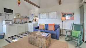 Ein gemütliches Wohnzimmer mit einem blauen Sofa, einem Holztisch, einem Schaukelstuhl und einer Küchenzeile mit weißen Schränken und einer Mikrowelle.