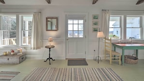 Ein Zimmer mit Holzbalkendecke, einer weißen Tür, einem Tisch mit Lampe, einem Stuhl und einem Fenster mit Vorhängen.