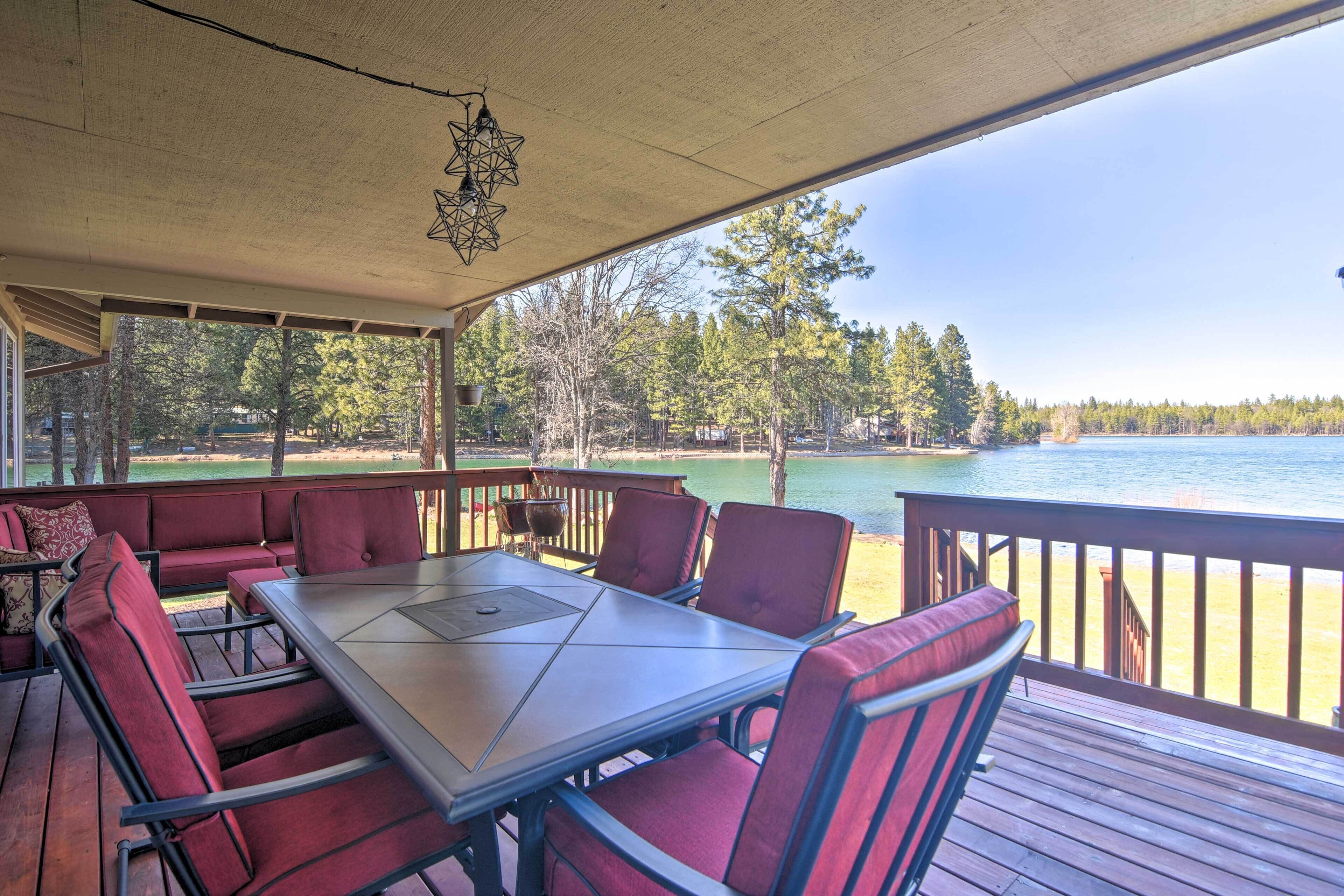 A covered outdoor seating area with a table and chairs, overlooking a lake and forest.